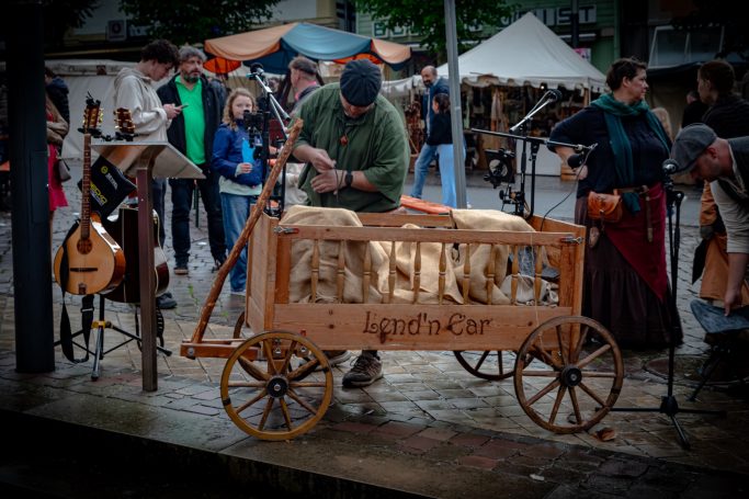 Ein Handwerker schnitzt Holzfiguren an einem Marktstand mit Zuschauern im Hintergrund.