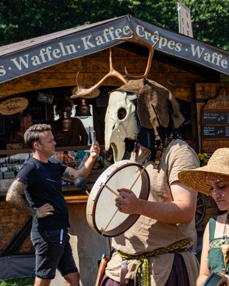 Person in einem Tierkopfkostüm spielt Trommel vor einem Verkaufsstand.