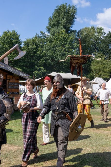 Menschen in historischer Kleidung parken auf einem Markt mit Zelten und Bäumen im Hintergrund.