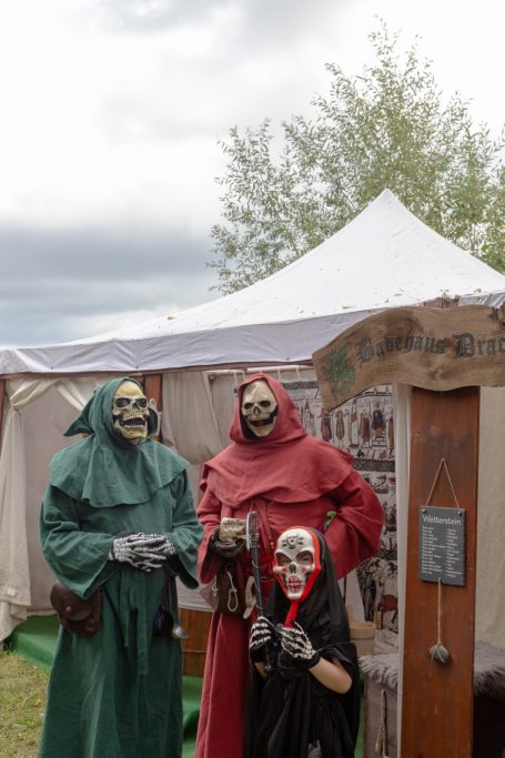 Drei Personen in Kostümen mit Totenkopf-Masken stehen vor einem Zelt.
