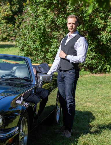 Mann im Anzug steht neben einem Cabrio auf einer grünen Wiese.