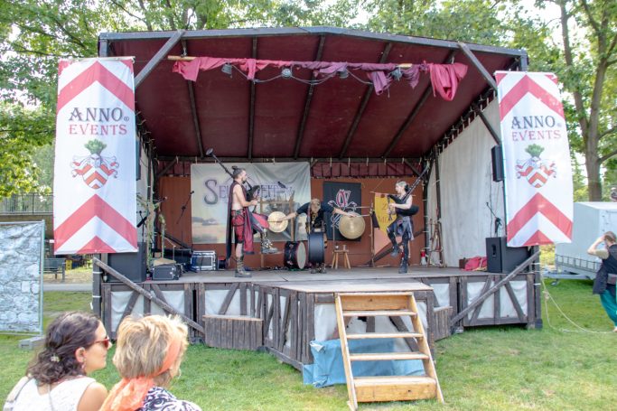 Musikgruppe tritt auf einer Bühne mit dekorativen Banner und Publikum im Vordergrund auf.