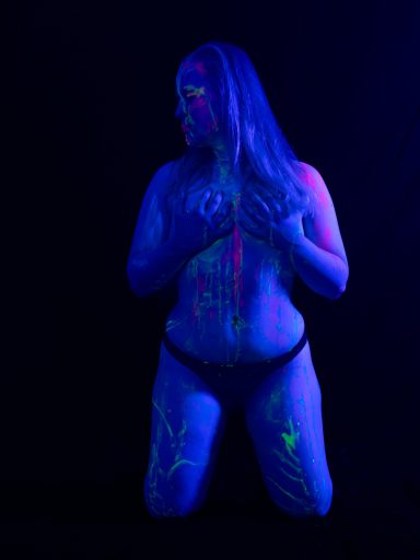 Eine Person mit leuchtenden Körperfarben, kniend vor einem dunklen Hintergrund.