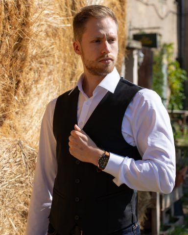 Mann in schwarzem Weste und weißem Hemd, vor einem Heuballen stehend.