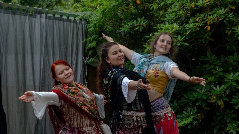 Drei Frauen in bunten Kostümen tanzen fröhlich im Freien vor grüner Kulisse.