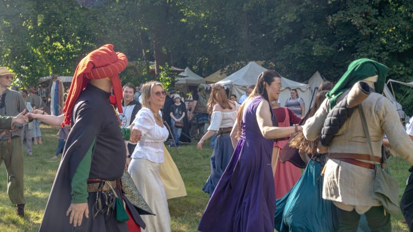 Personen in historischen Kostümen tanzen fröhlich auf einer Wiese bei einer Veranstaltung.