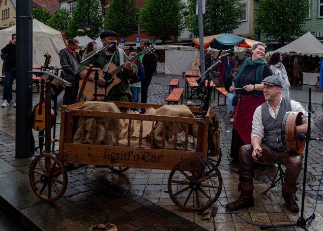 Marktbeschicker mit Schafen, Trachten und Musikinstrumenten in regnerischer Umgebung.