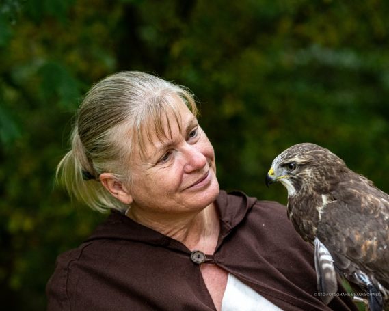 Frau mit langen blonden Haaren lächelt einen Greifvogel an, der auf ihrem Arm sitzt.