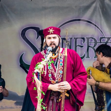 Mann in rotem Gewand mit Mikrophon, umgeben von Musiker auf einer Bühne.