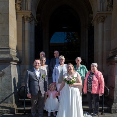 Hochzeitsgesellschaft vor einem historischen Gebäude, Braut in weißem Kleid mit Blumenstrauß.
