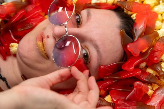 Person liegt mit rotem Gummibärchen und Popcorn umgeben, trägt runde Brille.