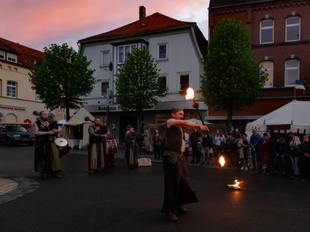 Feuerspuckender Akrobat in einer Stadt mit Bäumen und Zuschauern bei Sonnenuntergang.