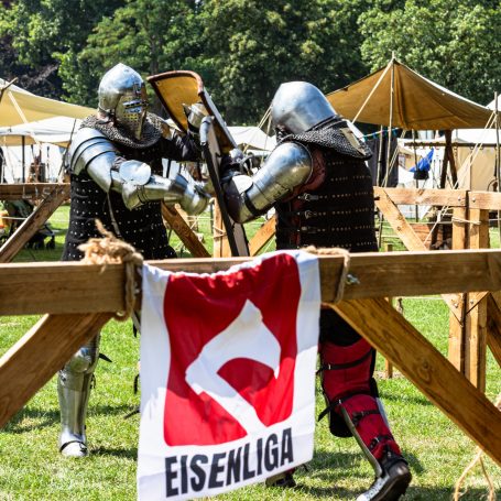 Zwei Ritter in Rüstung kämpfen in einem historischen Lager mit Zelten im Hintergrund.
