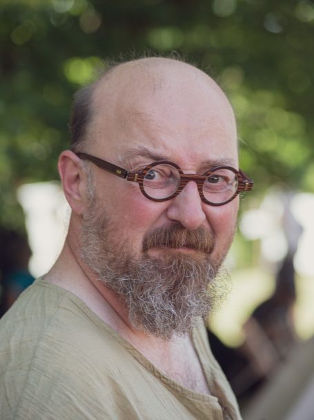 Ein Mann mit Brille und Bart, lächelnd, trägt ein einfaches, helles Oberteil.