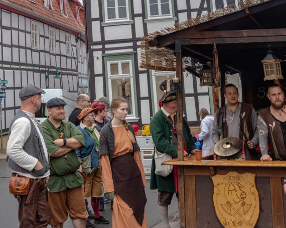 Marktfest mit kostümierten Personen, einem Stand und historischen Gebäuden im Hintergrund.