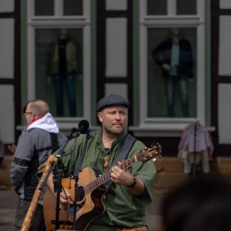 Mann mit Gitarre spielt Musik auf einer belebten Straße, im Hintergrund Schaufensterpuppen.