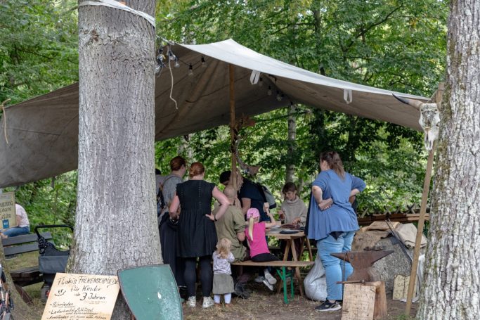 Menschen stehen unter einem Zeltdach und arbeiten an Holztischen im Wald.