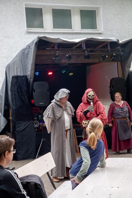 Zwei verkleidete Personen auf einer Bühne, eine Frau im Hintergrund beobachtet.