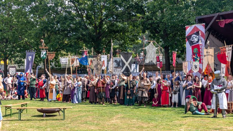 Eine Gruppe von Menschen in historischen Kostümen steht vor Bannern und einer Bühne im Freien.