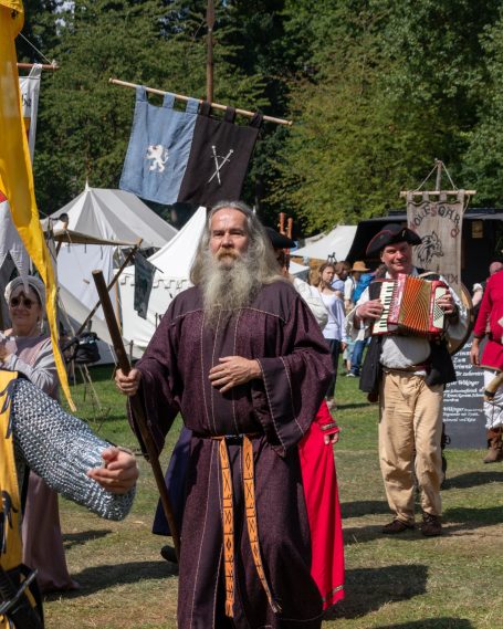 Mann in mittelalterlicher Kleidung mit Bart, umgeben von Festteilnehmern und Zelten.