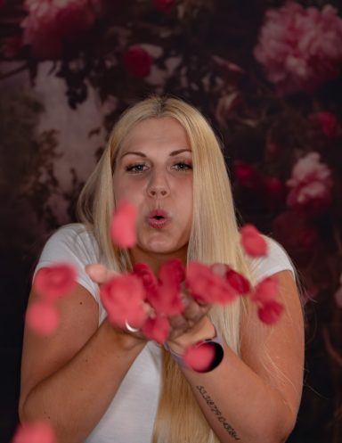 Frau mit langen Haaren bläst Rosenblätter in die Luft vor einem blumigen Hintergrund.