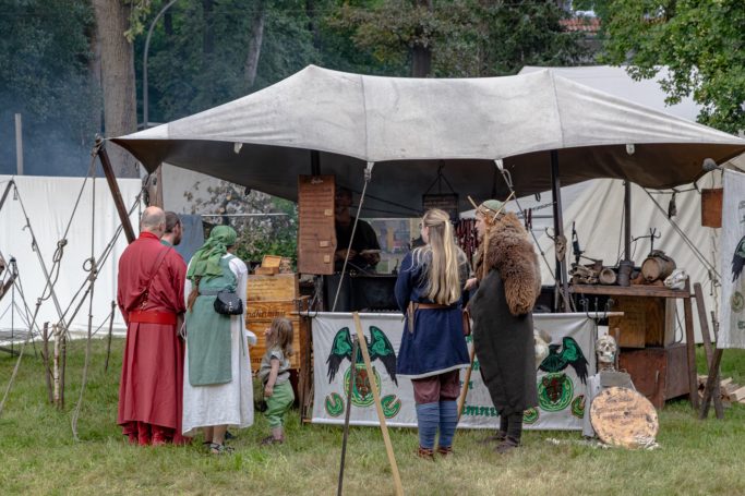 Mittelalterliche Lager-Szene mit kostümierten Personen vor einem Marktstand.