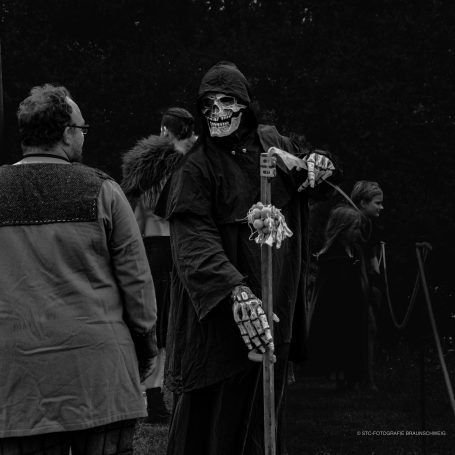 Ein Skelett in schwarzer Robe mit einem Staffel und einem Totenschädel im Hintergrund.