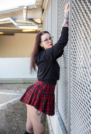 Frau in einem kurzen karierten Rock posiert an einer Gitterwand.