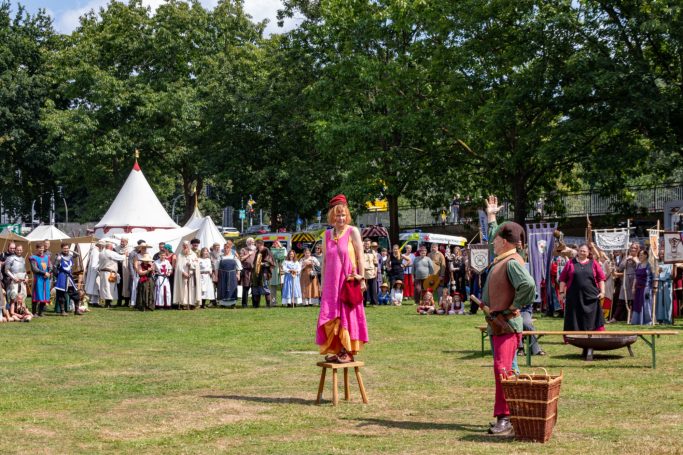 Zwei Akteure in Kostümen unterhalten eine Menschenmenge auf einer Wiese beim Fest.