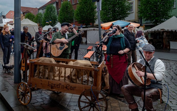 Mittelalterliche Musikgruppe mit Instrumenten und Schafen auf einem Stadtmarkt.