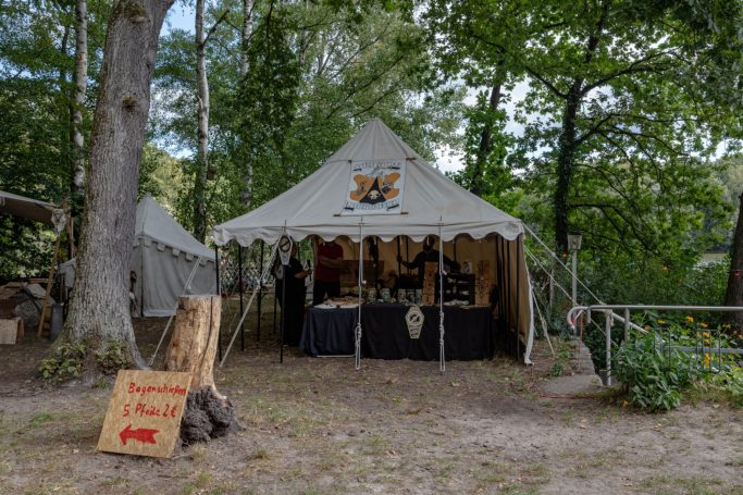 Markierte Zeltlager im Wald mit einem Verkaufsstand und einer Holztafel.
