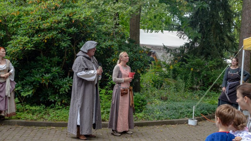 Zwei Personen in historischer Kleidung stehen in einem Garten, umgeben von Bäumen.