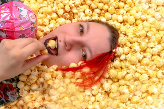 Frau mit roten Haaren, die in einem Haufen Popcorn liegt und Popcorn isst.