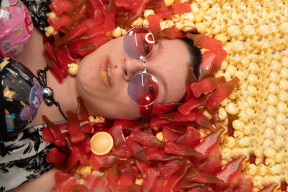 Frau liegt auf einem Bett aus Popcorn und buntem Süßigkeiten, mit Sonnenbrille und fröhlichem Gesicht.
