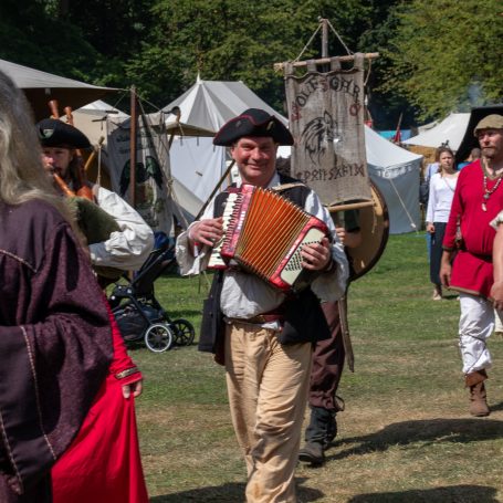 Ein Mann in historischer Kleidung spielt Akkordeon auf einem Fest. Hintergrund mit Zelten.