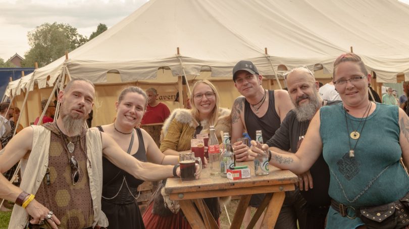 Gruppe von fünf Personen lächelt vor einem Zelt, Getränke in Händen, freundliche Atmosphäre.