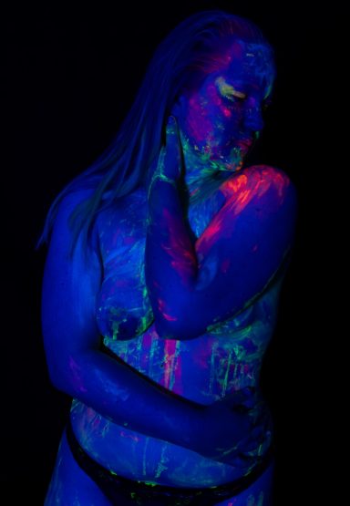 Eine Frau mit leuchtenden, bunten Körpermalereien steht vor dunklem Hintergrund.