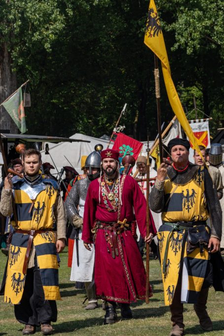 Personen in historischer Rüstung mit Bannern, im Freien, bei einem Fest oder Turnier.