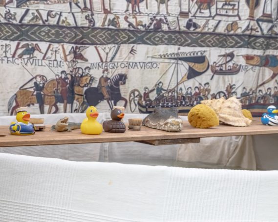 Bunte Gummienten und Muscheln auf einer Holzplatte, im Hintergrund historische Wandmalerei.