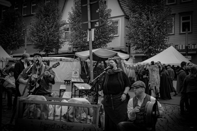 Musikerinnen und Musiker spielen auf einem Marktfest, umgeben von Besuchern und Ständen.