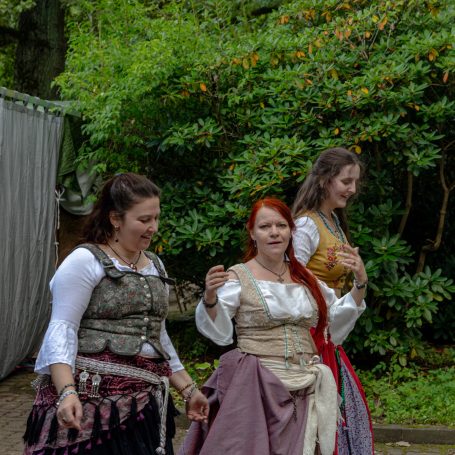 Drei Frauen in historischer Kleidung, die in einem Garten stehen und sich unterhalten.