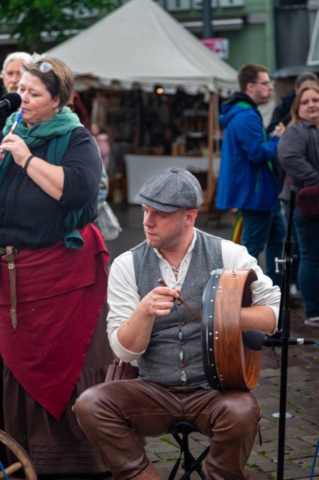 Frau mit Mikrofon steht neben einem Mann, der eine Trommel spielt, auf einem Markt.