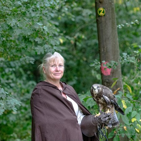 Frau mit einem Falken auf dem Arm steht vor einem Baum im Wald.