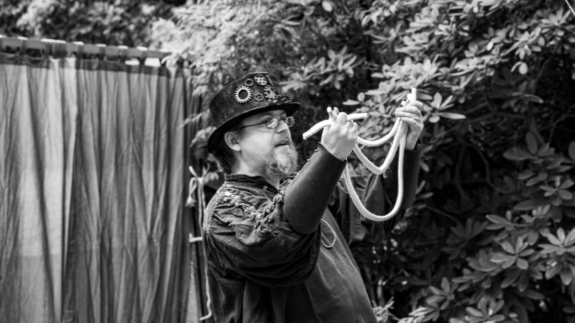 Ein Mann in schwarzer Kleidung hält Seile und steht in einem Garten.