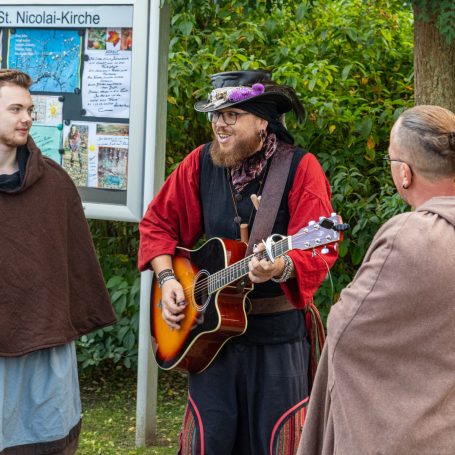 Drei Männer in historischen Kostümen unterhalten sich, einer spielt Gitarre.