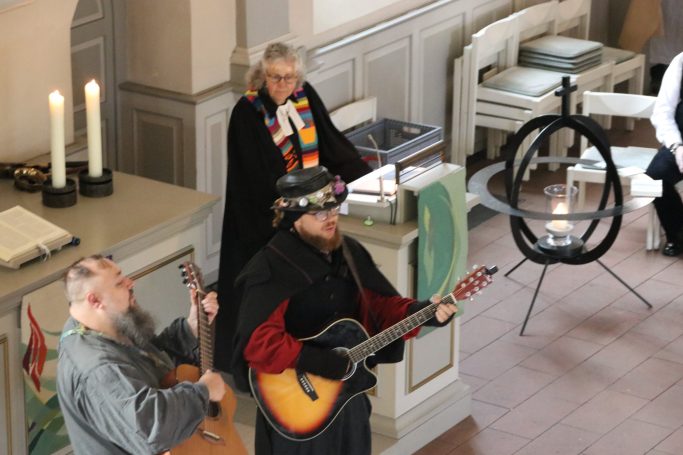 Musiker mit Gitarre, während eine Person im Hintergrund spricht, in einer Kirche.