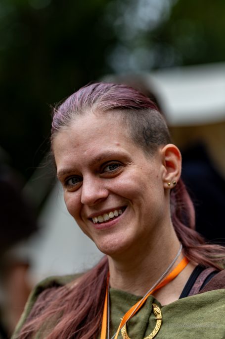 Frau mit lila Haar und Ohrring lächelt freundlich, trägt ein grünes Oberteil.