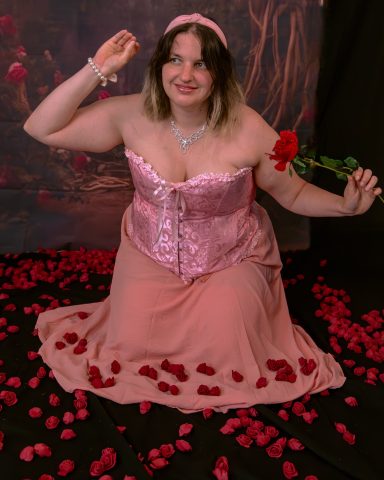 Frau in pinkem Kleid, sitzt auf Rosenblättern und hält eine rote Rose in der Hand.