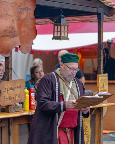 Mann in historischer Kleidung liest von einem Manuskript an einem Marktstand.