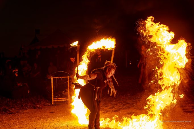 Person jongliert mit brennenden Fackeln vor dunklem Hintergrund. Flammen leuchten intensiv.